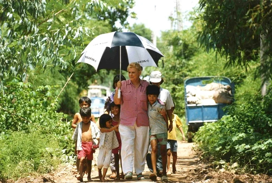 Christina Noble under an umbrella with a crowd of children on a dirt track, 1990s