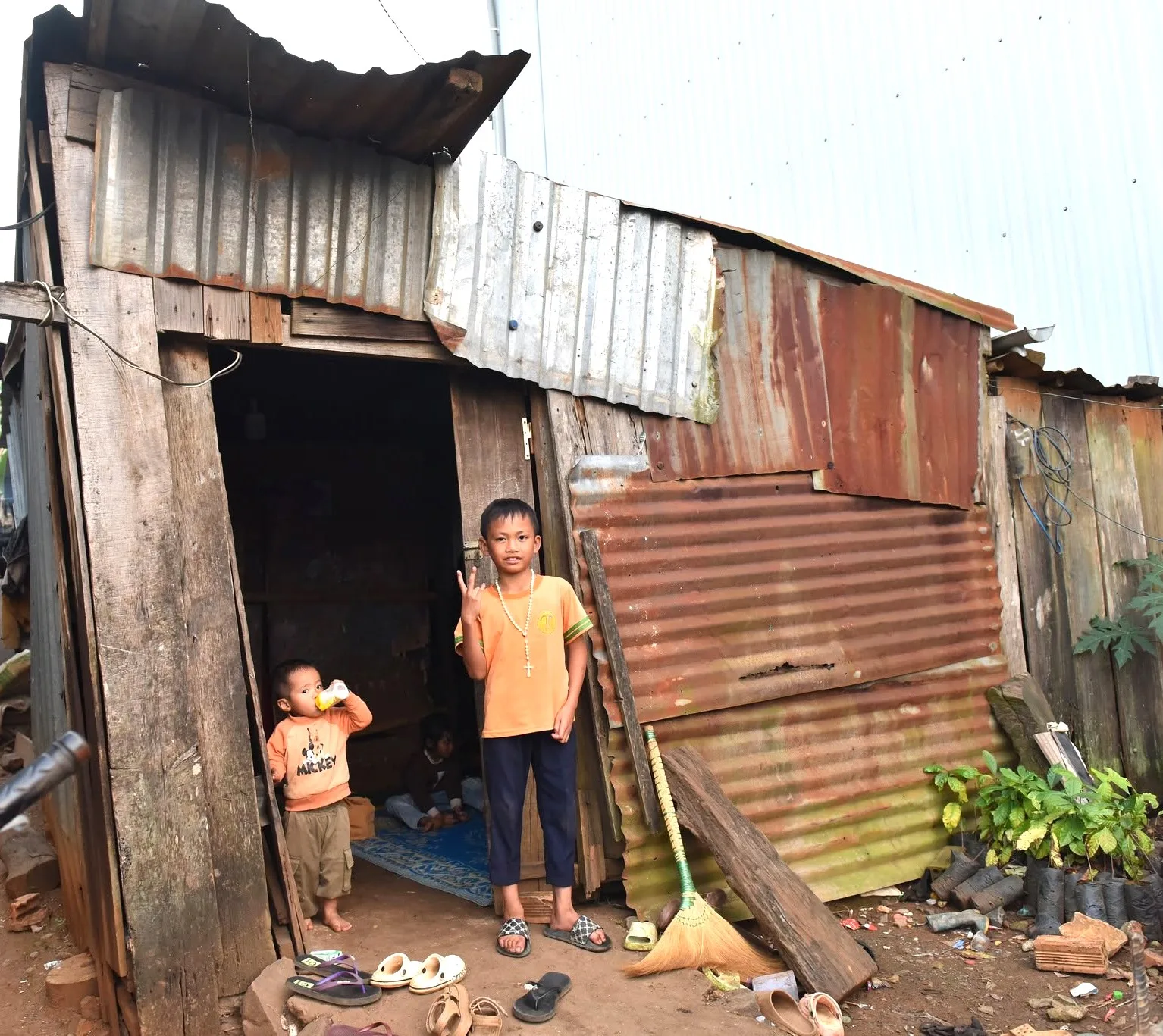 A boy in an orange shirt makes a peace sign outside his home, his younger brother behind him