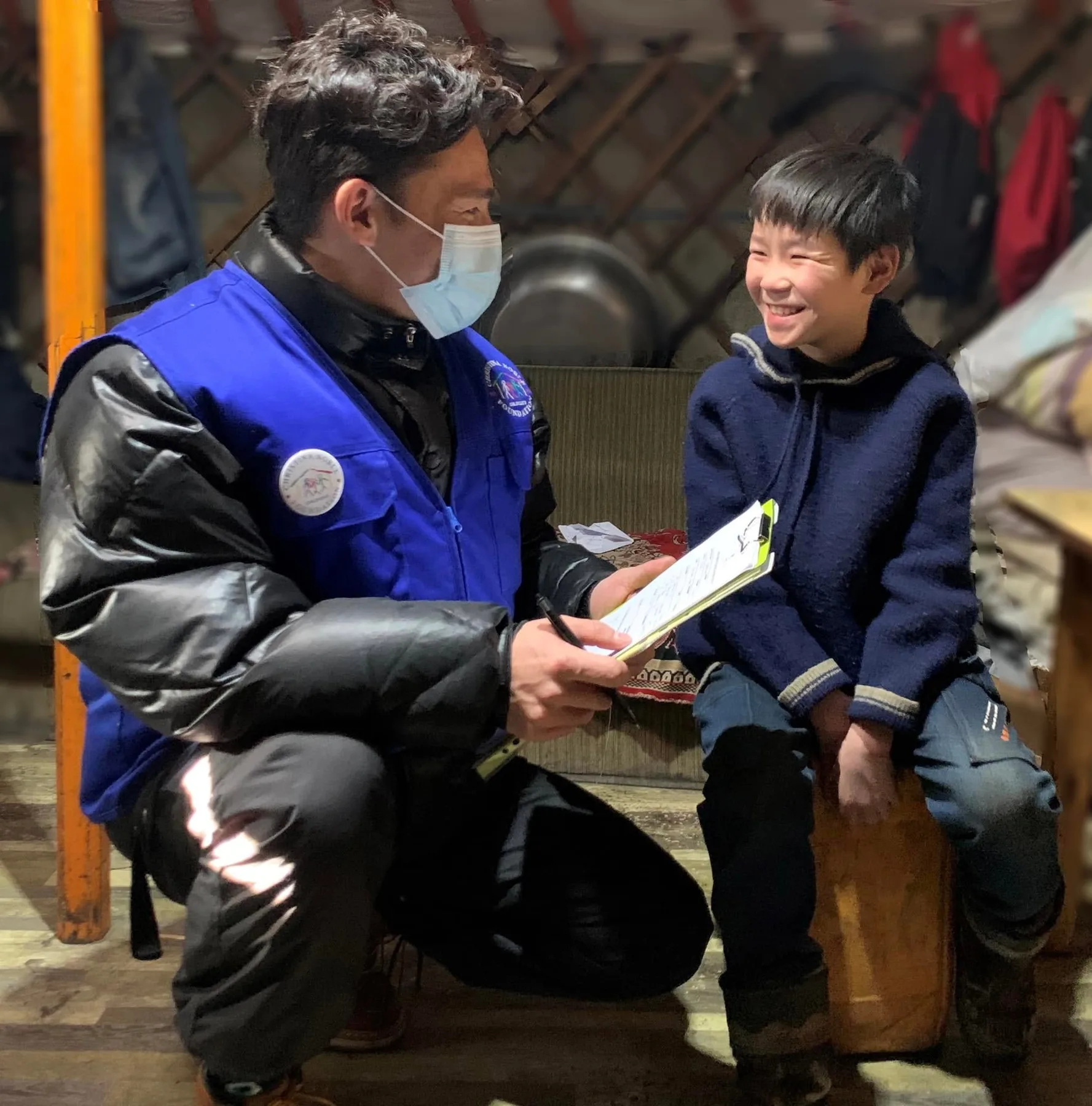 A CNCF social worker in a blue vest laughs with a boy inside a ger